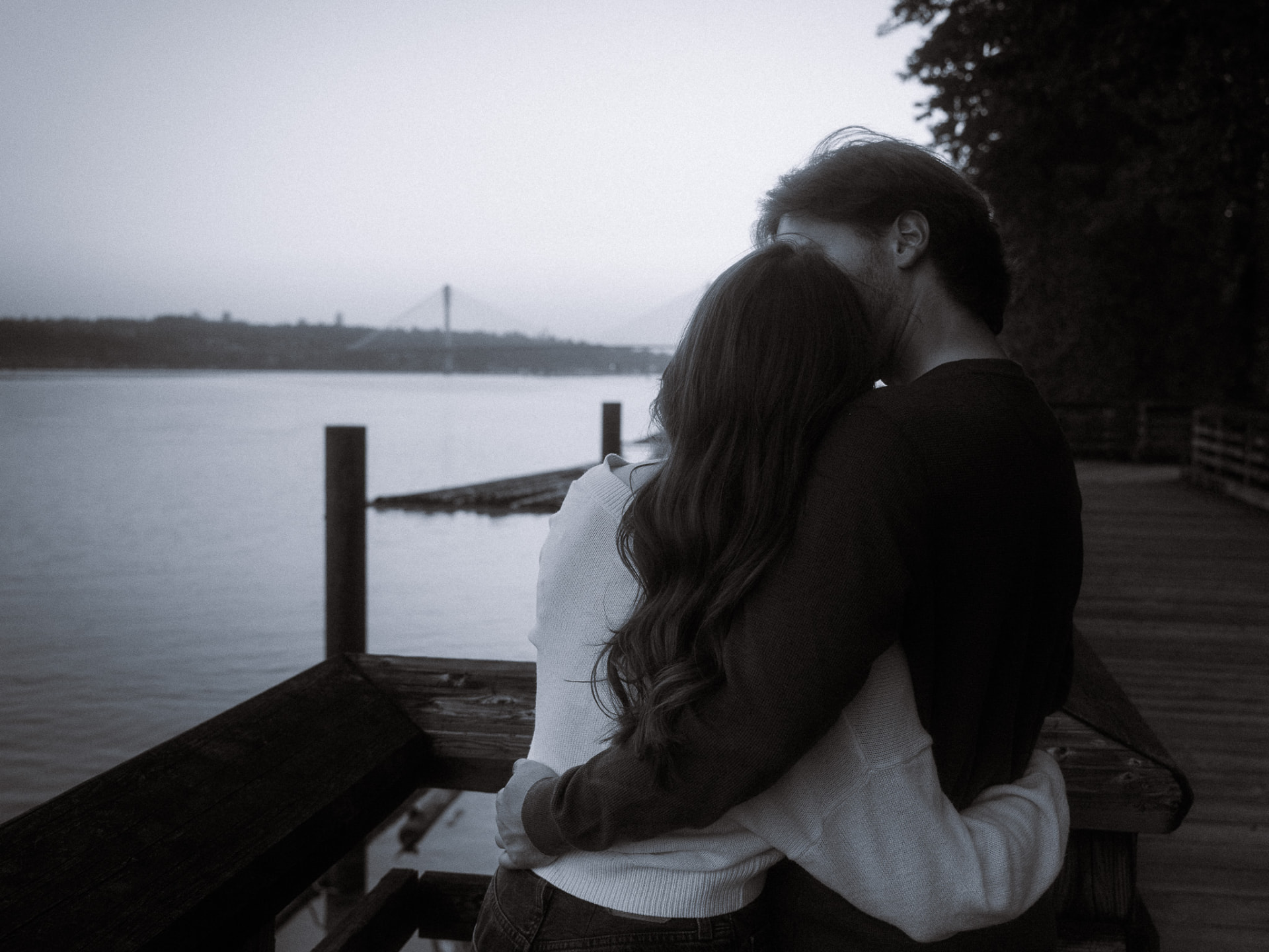 Black and white photo of a couple in a tender embrace on a wooden boardwalk overlooking the water at dusk in Metro Vancouver