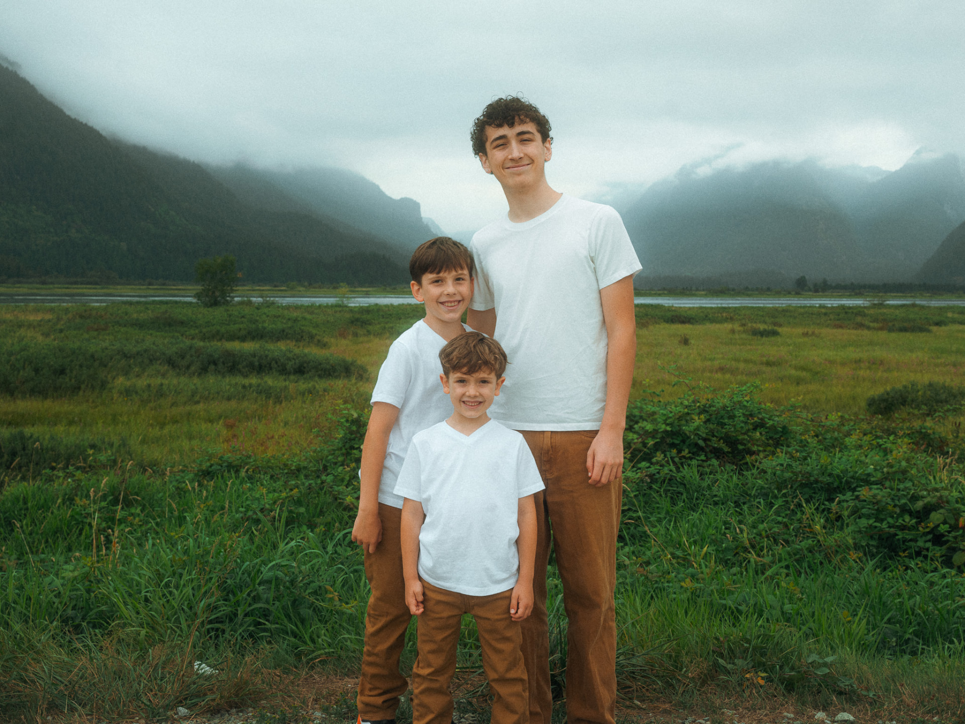 Three brothers smiling together in tall grass with fog-covered mountains behind them near Metro Vancouver