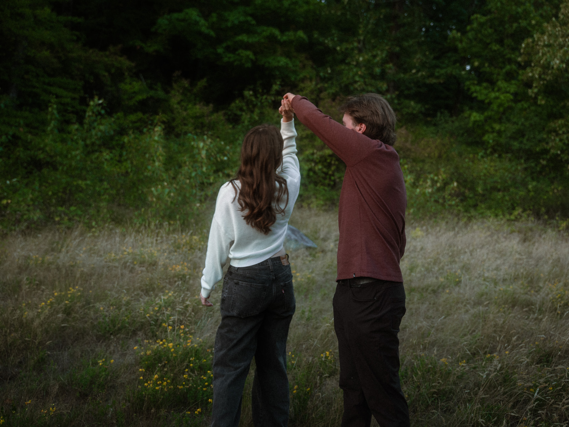 Couple seen from behind holding hands while dancing in a grassy field near a Metro Vancouver forest