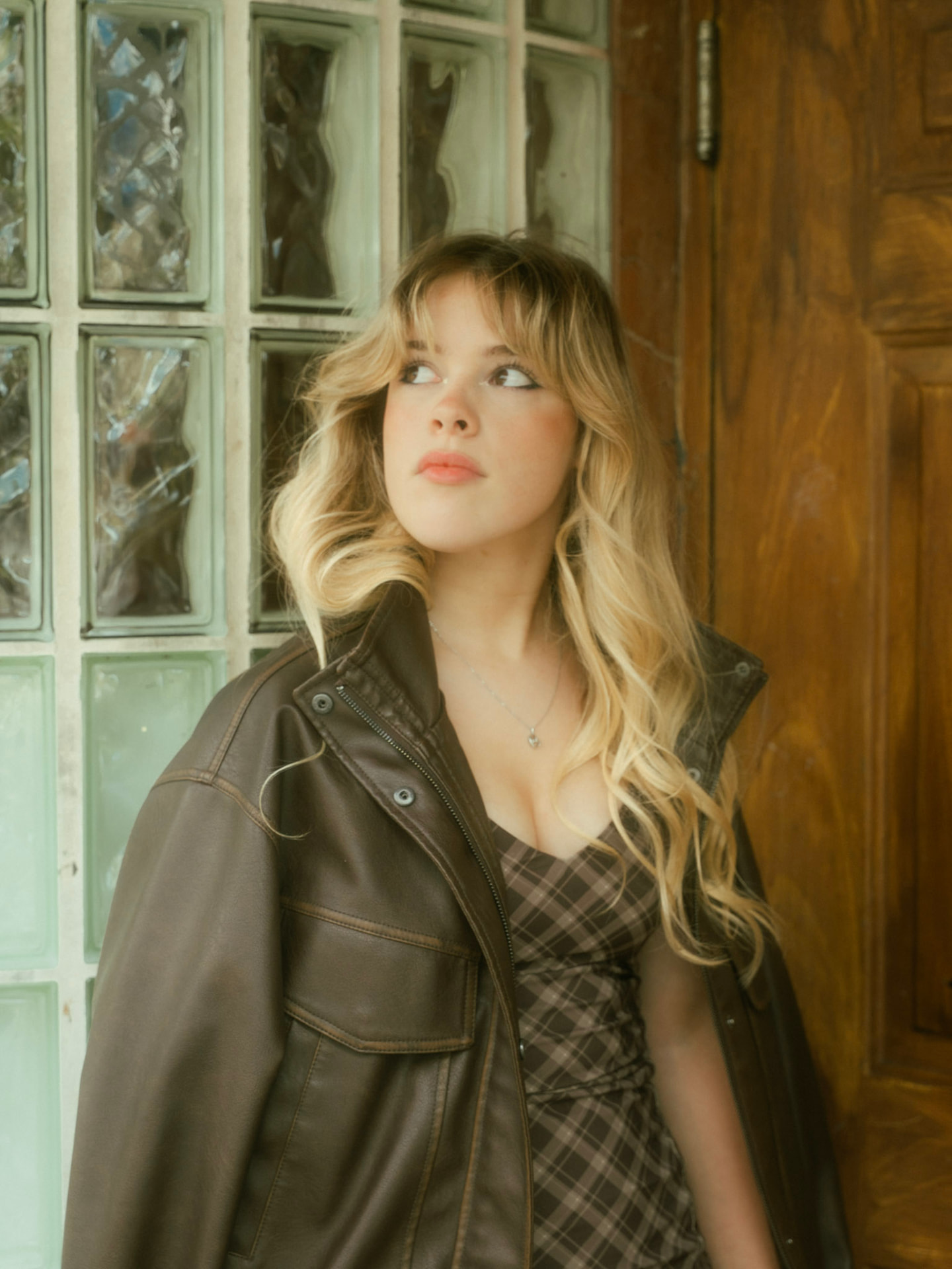 Close-up of a woman in a plaid dress and leather jacket in a pensive pose beside a vintage door in Vancouver