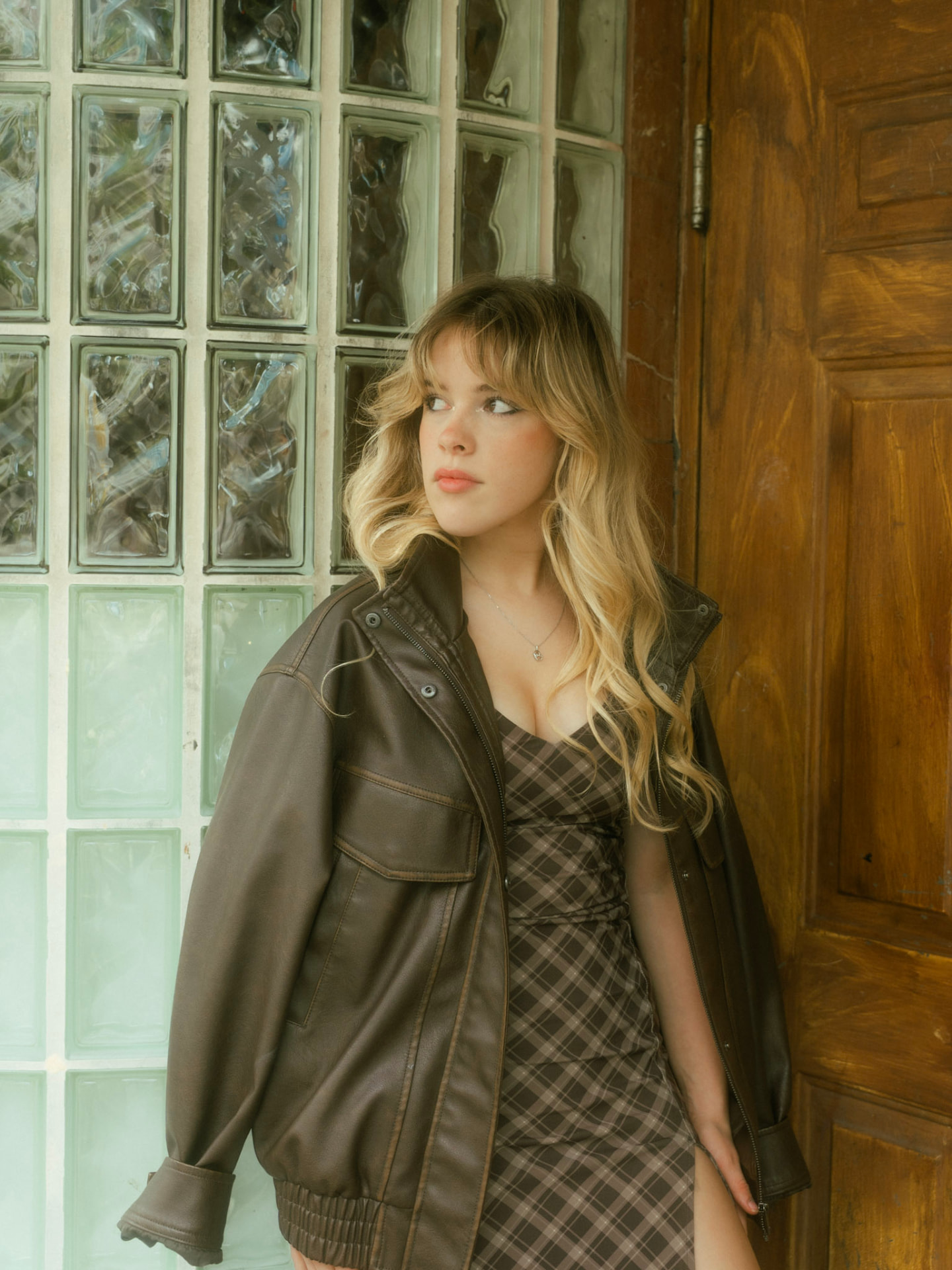 Woman in a plaid dress with a leather jacket draped over her shoulders standing by a glass block wall in Vancouver