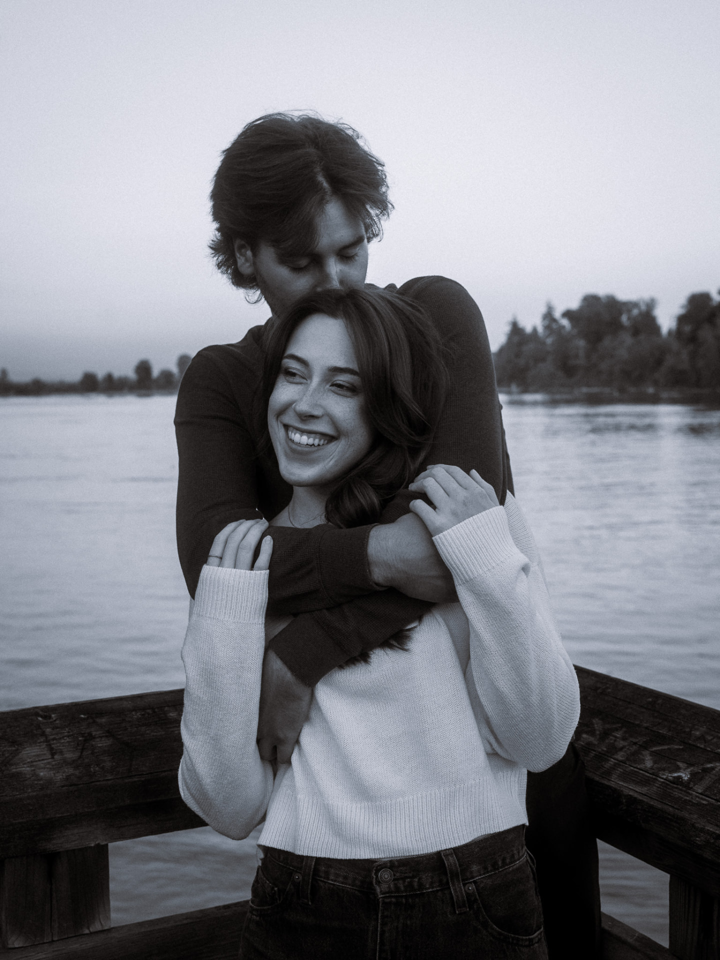 Black and white photo of a man hugging a woman from behind and kissing her forehead on a riverside dock in Metro Vancouver