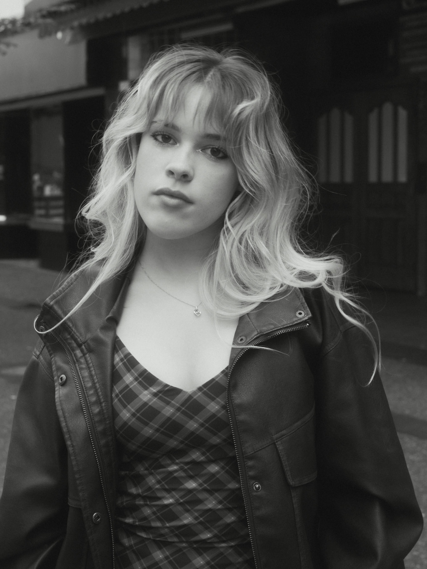 Black and white portrait of a woman in a plaid dress and leather jacket facing the camera on a Metro Vancouver street