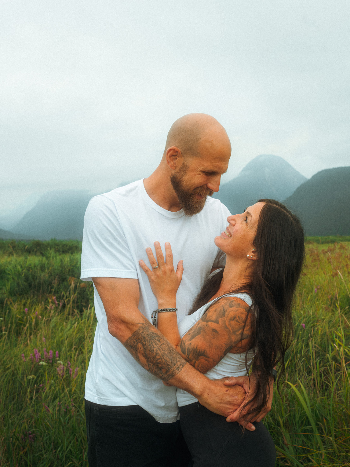 Mother and father sharing a romantic embrace in a grassy field with misty mountains behind them near Metro Vancouver
