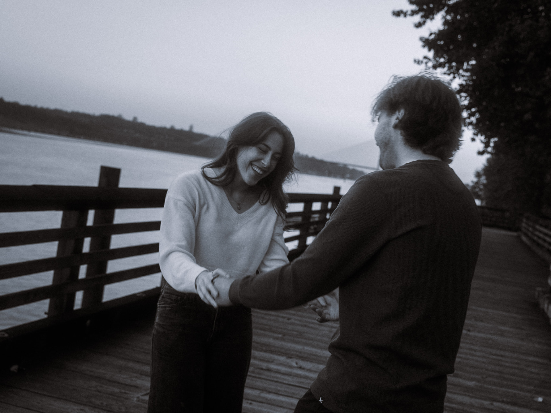 Black and white photo of a couple holding hands and laughing on a riverside boardwalk in Metro Vancouver