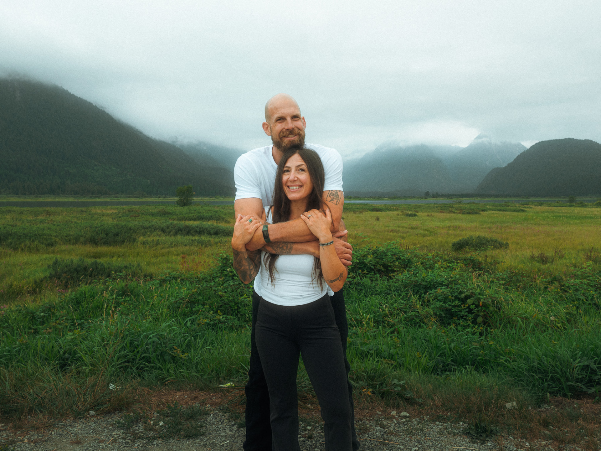 Mother and father embracing happily in a grassy landscape with foggy mountains in the distance near Metro Vancouver