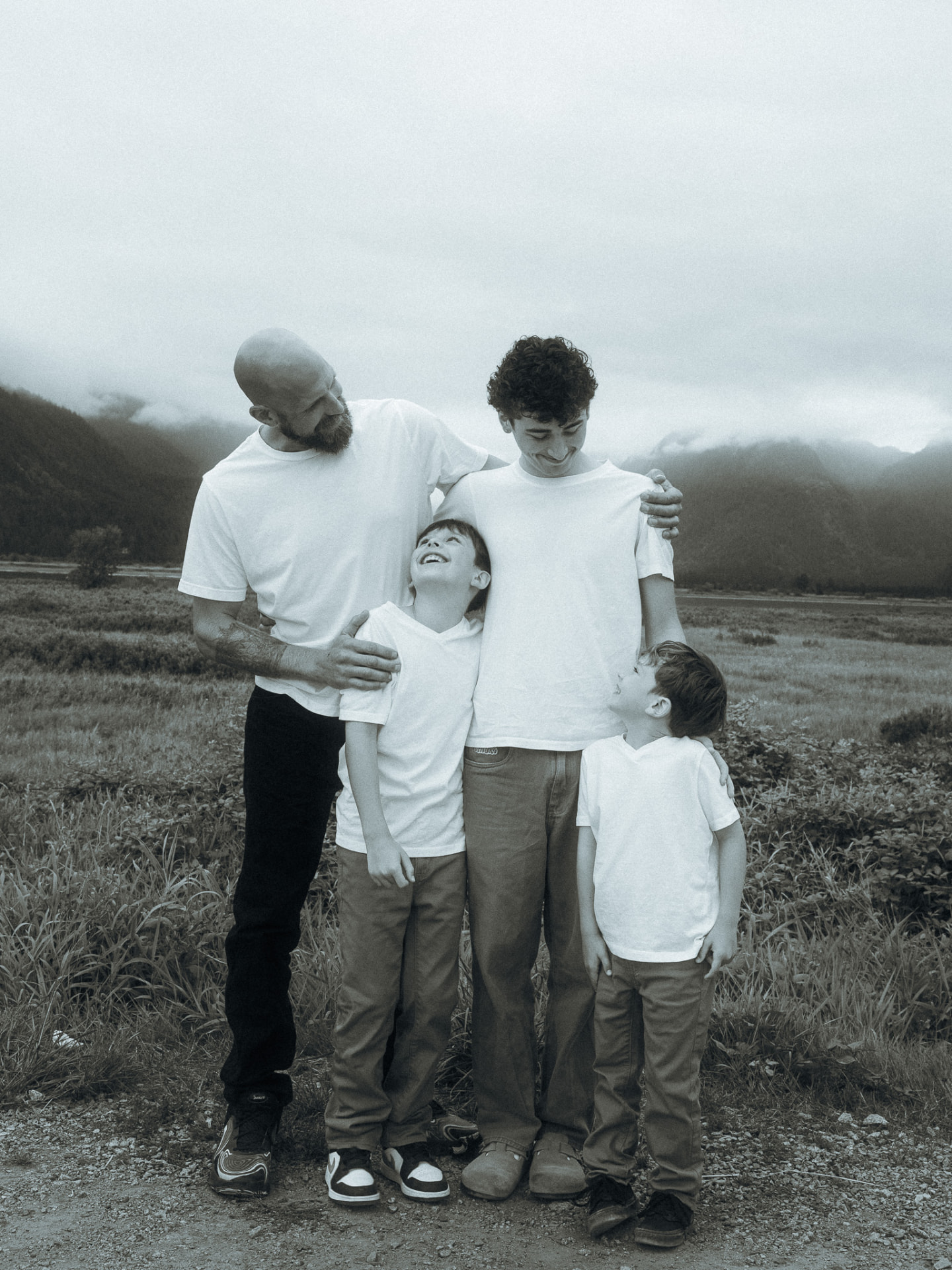 Black and white photo of a father and his three sons laughing together in a mountain-rimmed field near Metro Vancouver